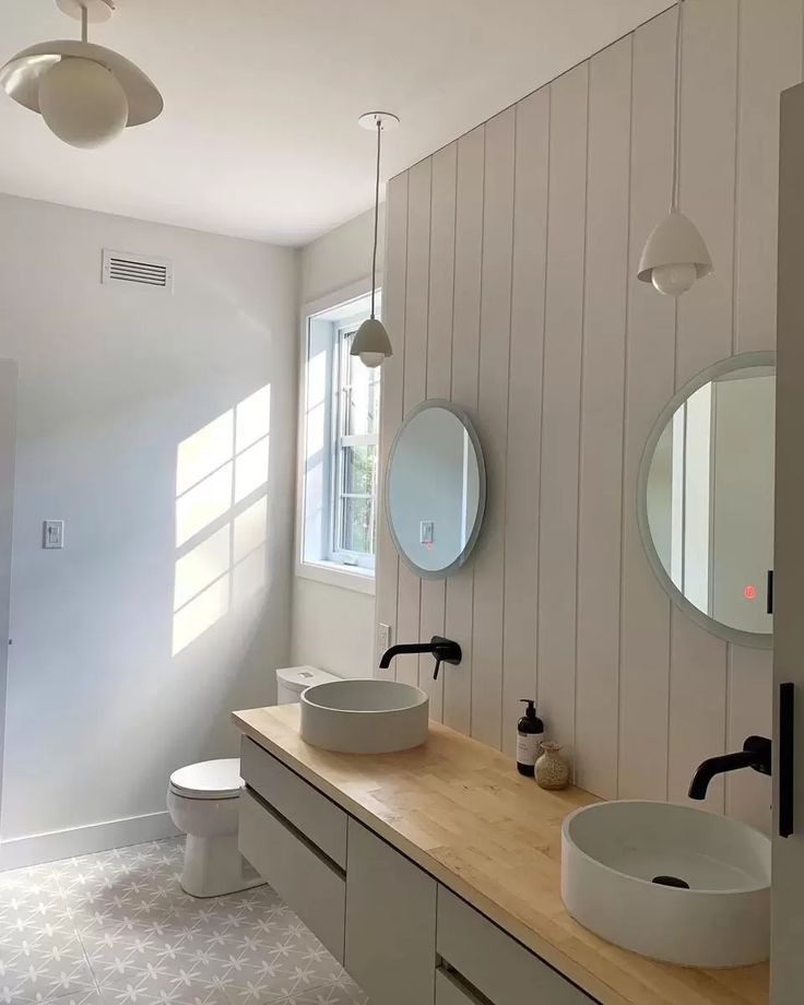 Minimalist bathroom with a wooden countertop and two vessel sinks. Round mirrors hang above the sinks. Light streams through a window. Calm ambiance.