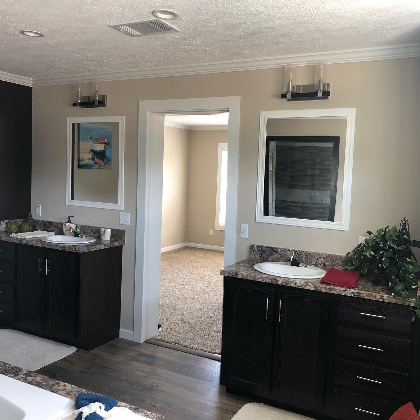 Modern bathroom with dark wood vanities and marble countertops. Two sinks, framed mirrors, and wall art above. A doorway leads to a carpeted room.
