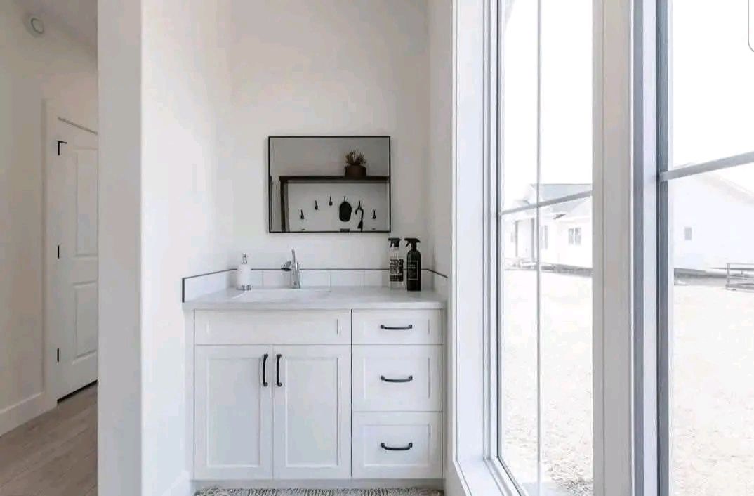 Minimalist bathroom with a white vanity, black handles, and a large mirror. Bright light streams through a big window, creating an airy feel.