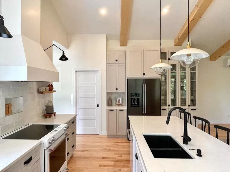 Modern kitchen with light wood flooring, a large white island, black fixtures, stainless steel appliances, and exposed wooden ceiling beams. Calm ambiance.