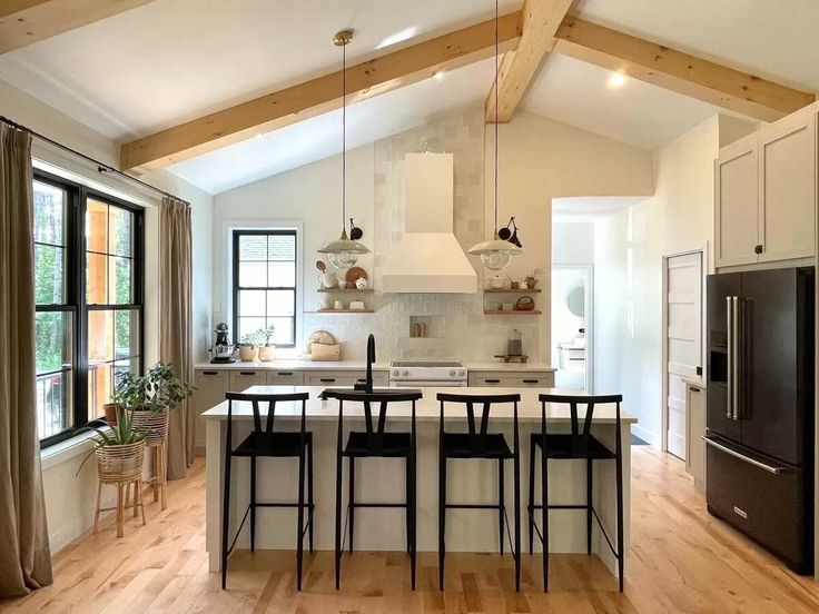 Bright kitchen with vaulted ceiling and wooden beams. Features include a white island with black stools, pendant lights, and large windows with curtains. Cozy and modern.