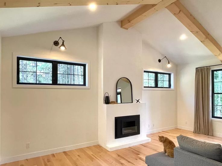A minimalist living room with a white fireplace and a tall mirror above it. Large windows showcase a forest view. A ginger cat sits on a gray couch, creating a cozy atmosphere.