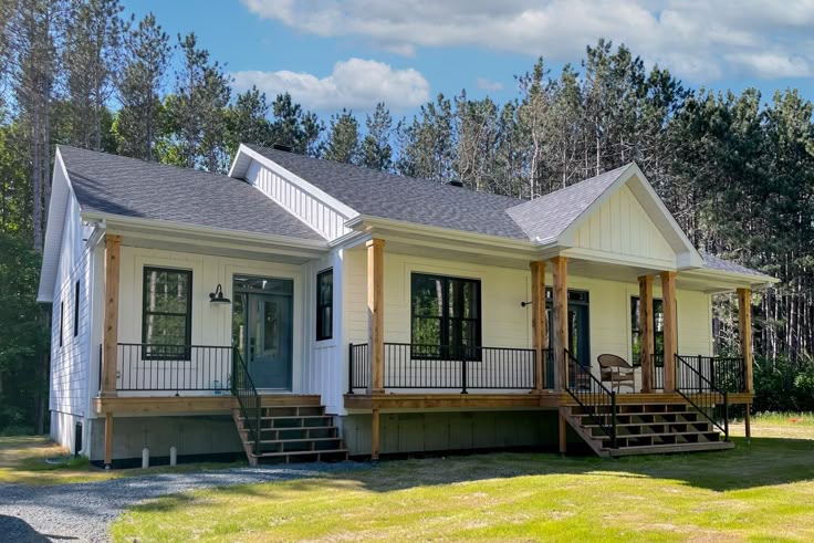 A modern white house with a dark roof, wooden pillars, and a spacious front porch. It sits on a lush lawn, framed by tall, dense trees under a blue sky.
