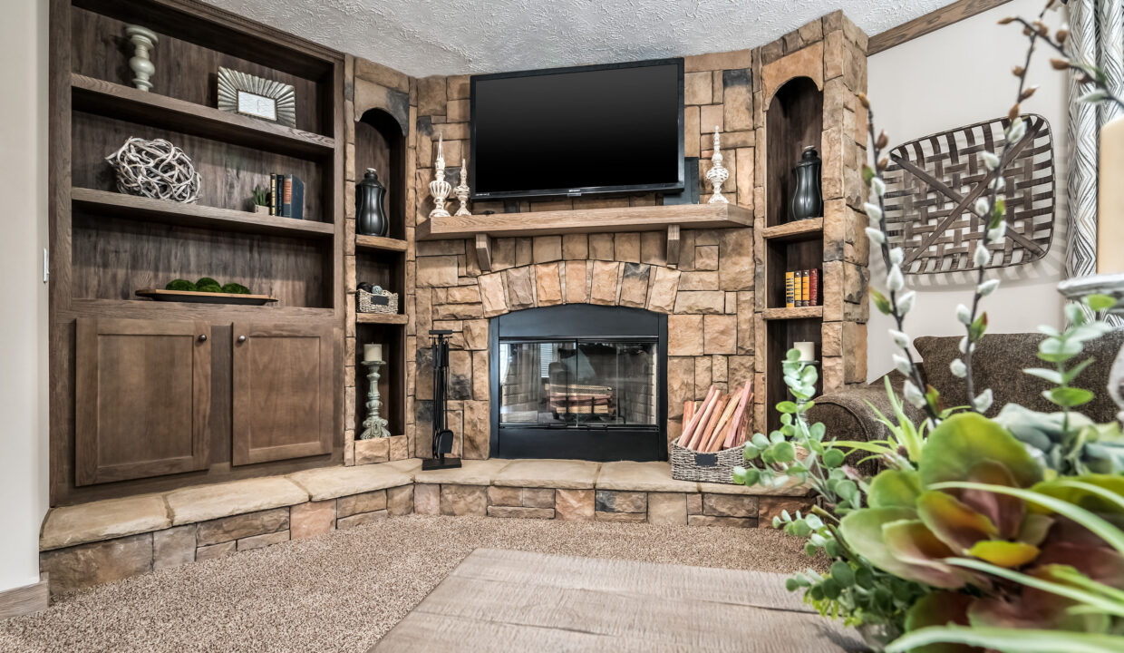 Cozy living room with a stone fireplace, shelves filled with decor, and a TV mounted above. A basket of firewood and green plants add warmth.