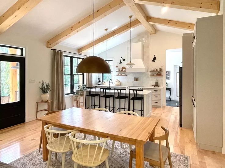 Spacious, bright kitchen-dining area with wooden beams, white walls, and light wood flooring. Features a wooden table, chairs, large pendant light, and modern kitchen island with black stools. Cozy and inviting ambiance.