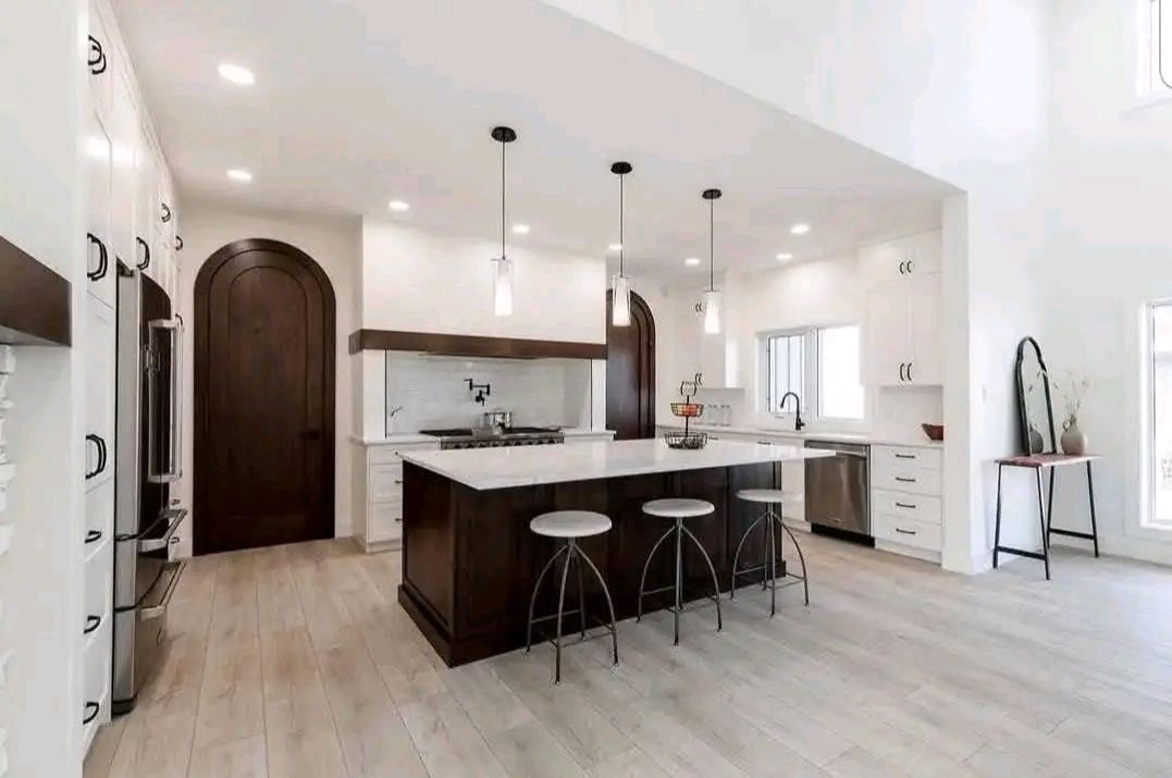 Spacious modern kitchen with white walls, wooden floors, and dark wood accents. Central island with pendant lights and three stools exudes elegance.