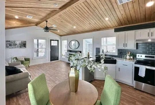 Modern open-plan living space with a wooden ceiling, light wood flooring, and a cozy kitchen. Green chairs and a round table with a tall vase highlight the dining area. Comfortable sofa in the living area adds warmth.