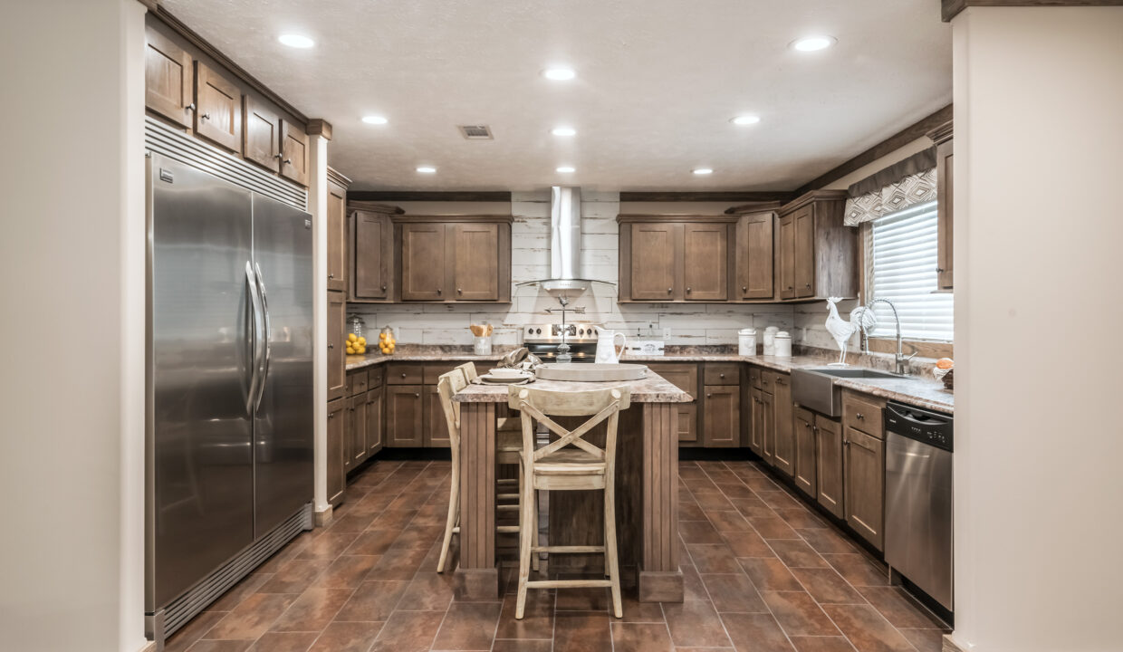 Spacious kitchen with rustic wood cabinetry, central island with stools, stainless steel appliances, and warm tile flooring under soft lighting.