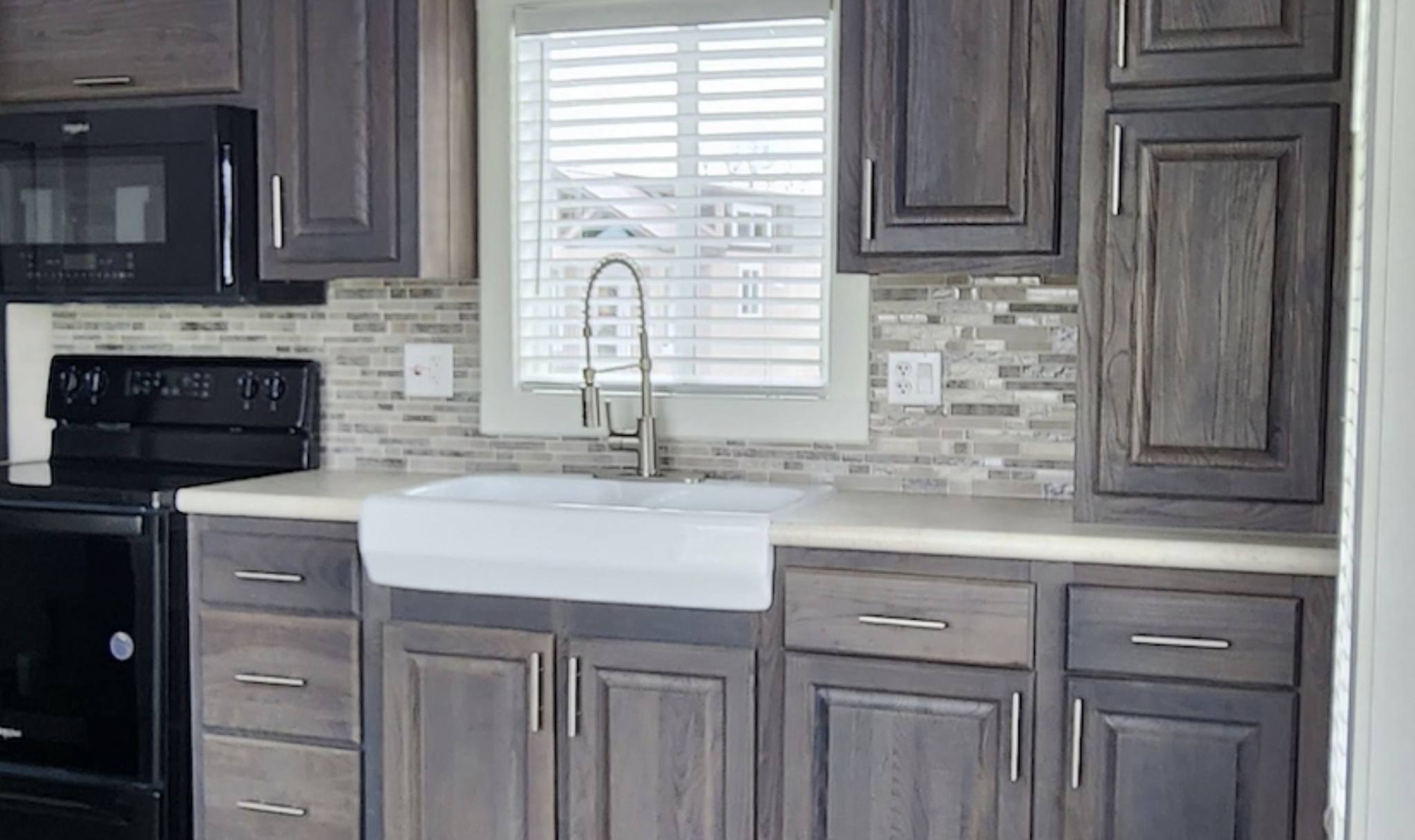  A close-up of a kitchen area featuring dark wood cabinets with silver bar handles and a large white farmhouse sink. Above the sink is a high-arc, industrial-style metal faucet and a window with white horizontal blinds. The backsplash consists of a neutral-toned horizontal mosaic tile, and black appliances are partially visible on the left.