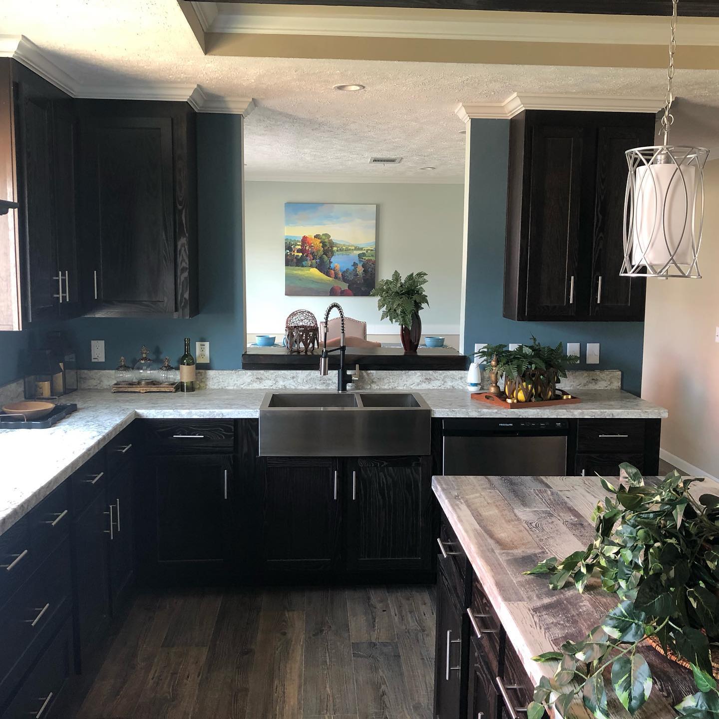 Modern kitchen with dark wood cabinets, marble countertops, and a stainless steel sink. A pendant light hangs above a wooden island with plants. A scenic painting and greenery create a serene, inviting atmosphere.