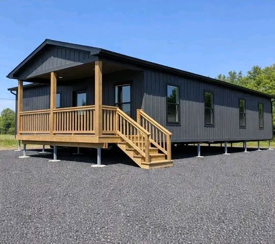 A dark gray modular home stands on a gravel lot, elevated on pillars. It features a small wooden porch with stairs, under a clear blue sky.