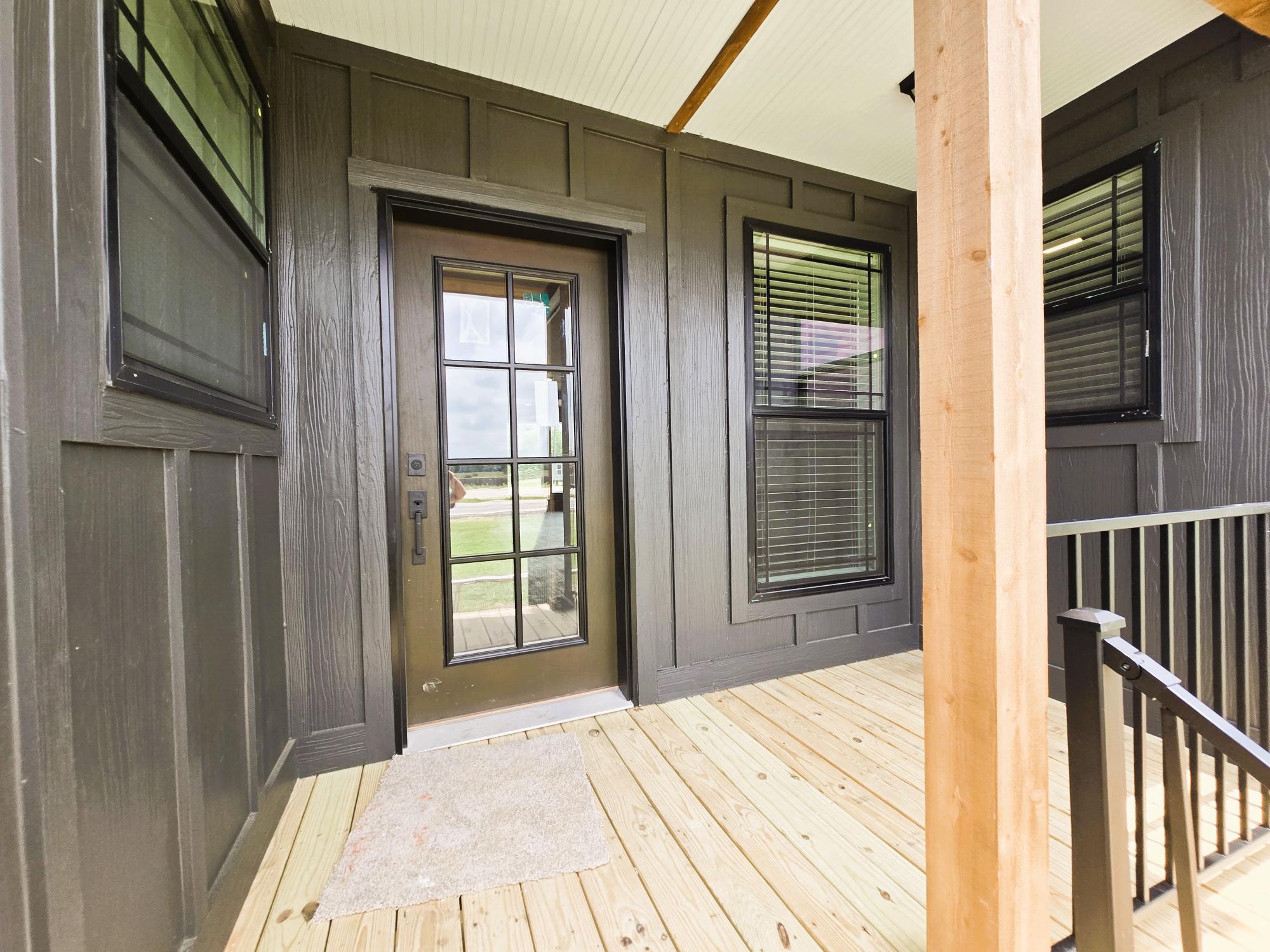 A welcoming porch with wooden flooring features a glass-paneled door flanked by windows with blinds. The exterior is dark-paneled, conveying a modern, rustic feel.