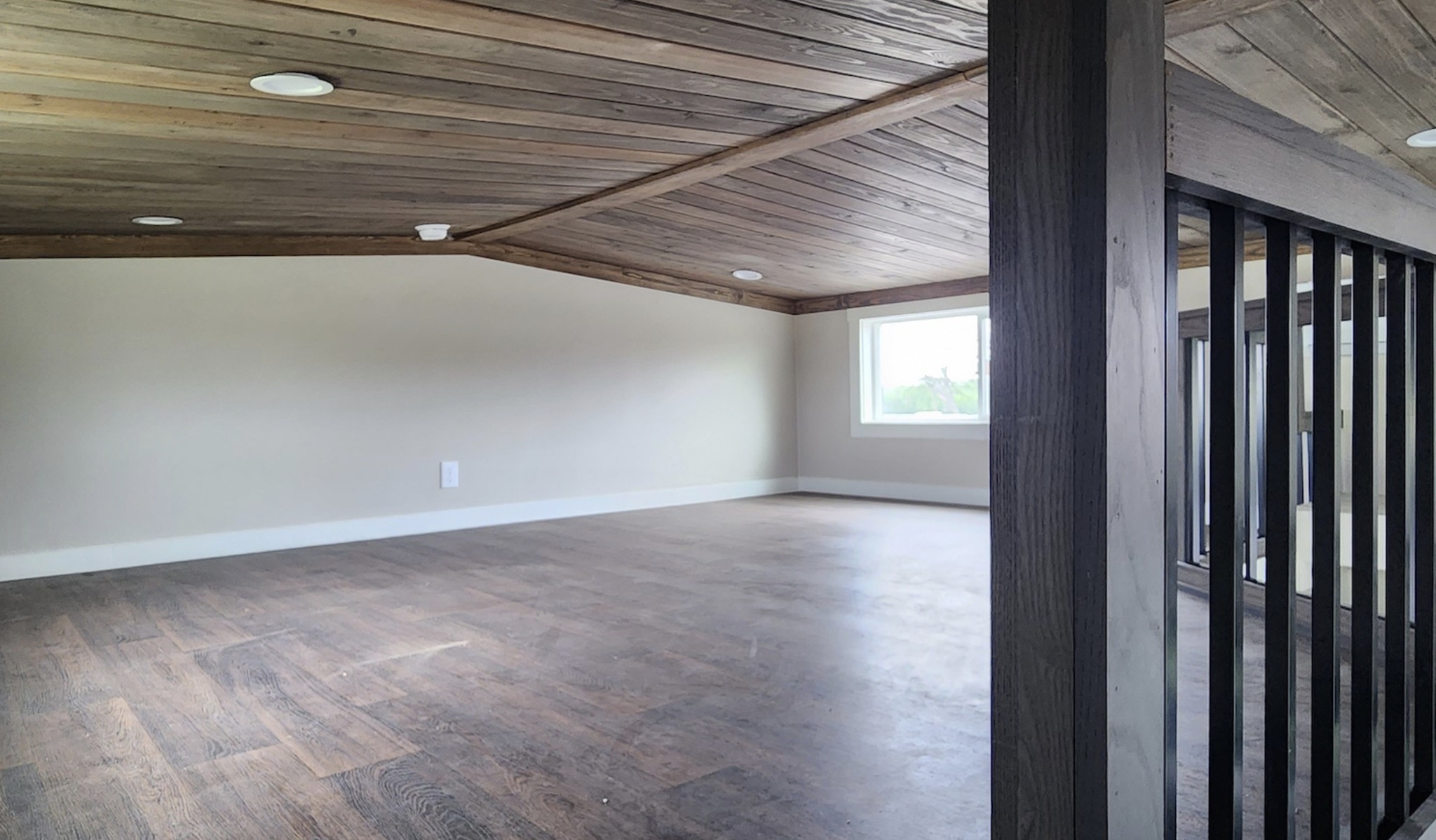  An empty loft area featuring a dark wood planked ceiling with recessed lighting, light beige walls, and dark wood-look flooring. A small window sits on the far wall, and a dark wood railing with black metal spindles is visible on the right.