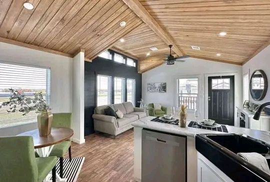 Modern open-concept living area with a wood-paneled ceiling. A gray sofa and two green chairs face a small table and a kitchen island. Bright natural light through large windows creates a welcoming atmosphere.