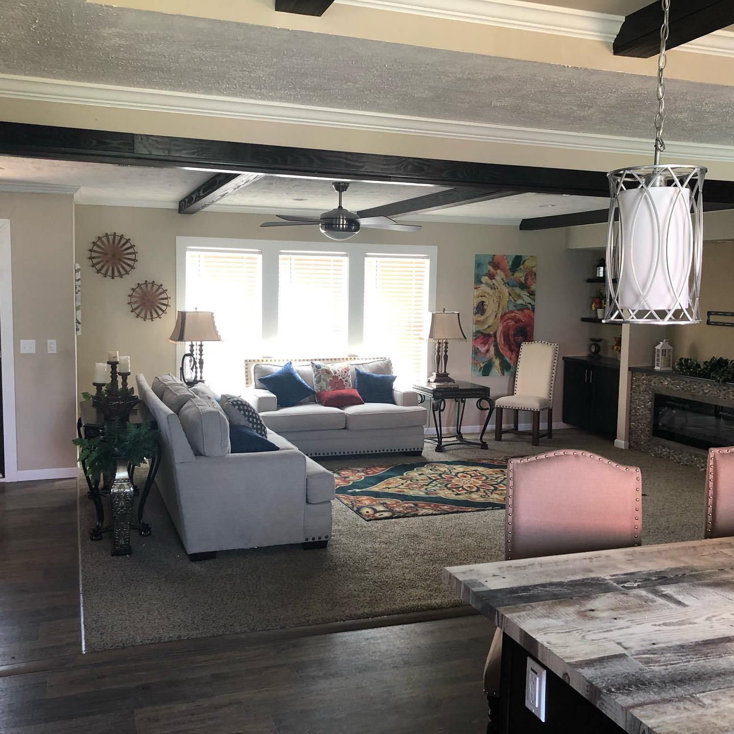 Spacious living room with a gray sofa, colorful cushions, and a floral rug. Features a modern chandelier, ceiling fan, and cozy fireplace. Natural light fills the room.
