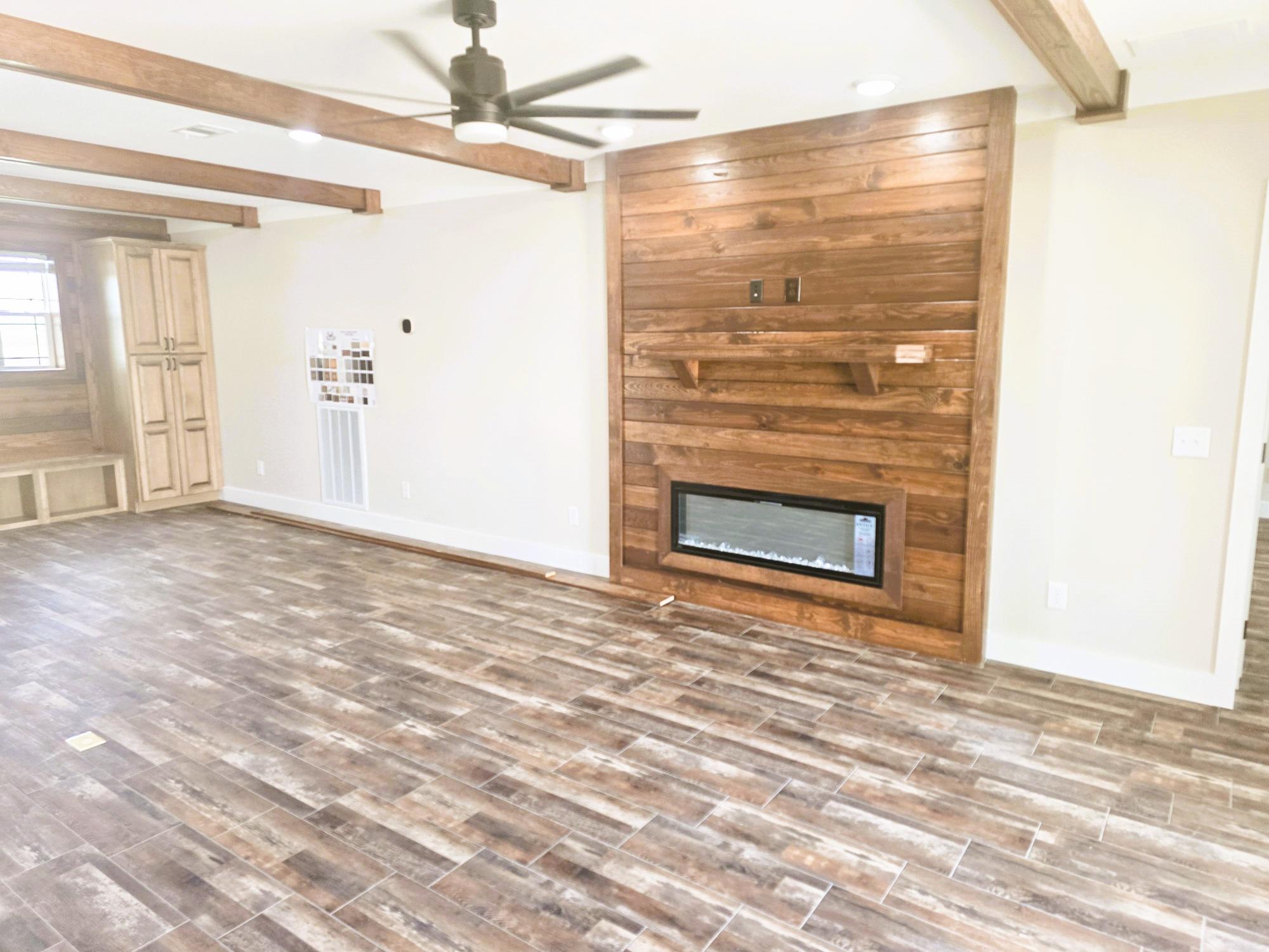 A modern living room with wood accents, featuring a wooden floor, beamed ceiling, and a wooden wall with a built-in electric fireplace and shelves. A ceiling fan and neutral-toned walls add to the contemporary feel.