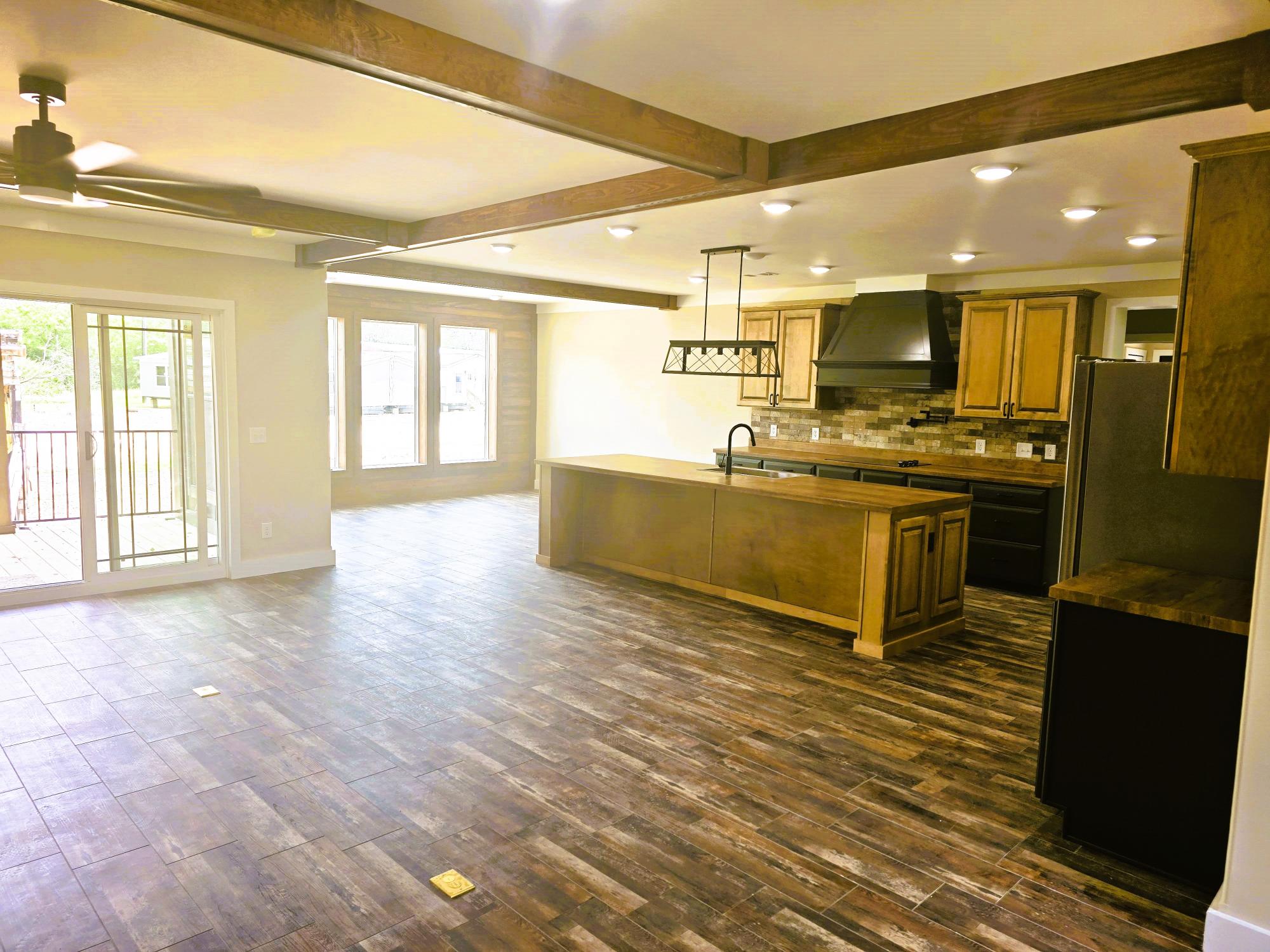 Spacious kitchen with dark wood flooring, a large island with sink, wooden cabinets, and a modern light fixture. Sliding door and large windows add brightness.