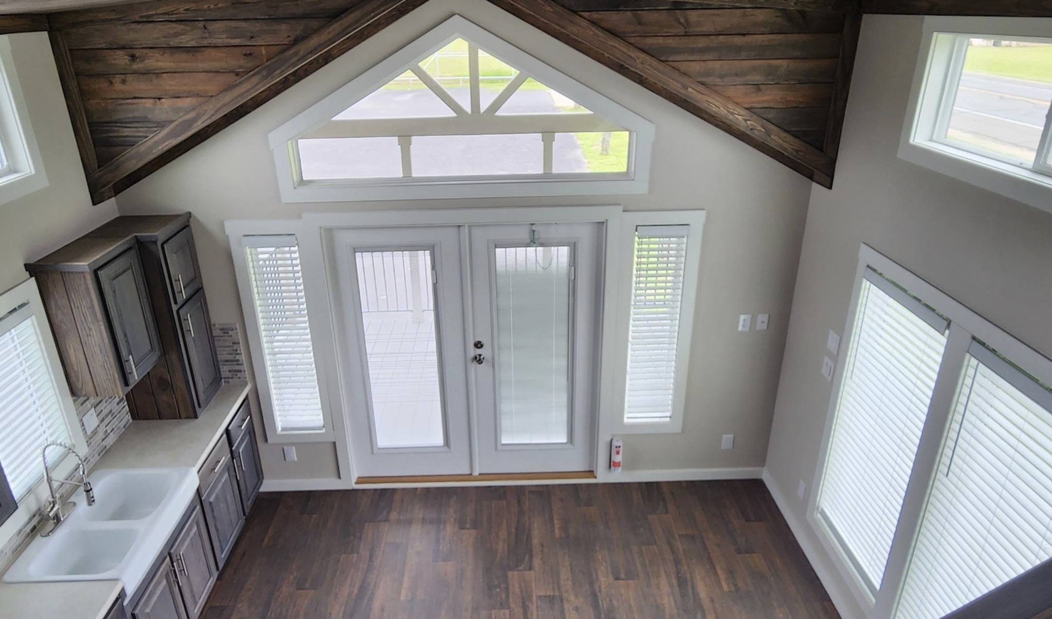 View of a bright, rustic kitchen with dark wood cabinets, white countertops, and a double sink. Sunlit French doors and large windows illuminate the space.