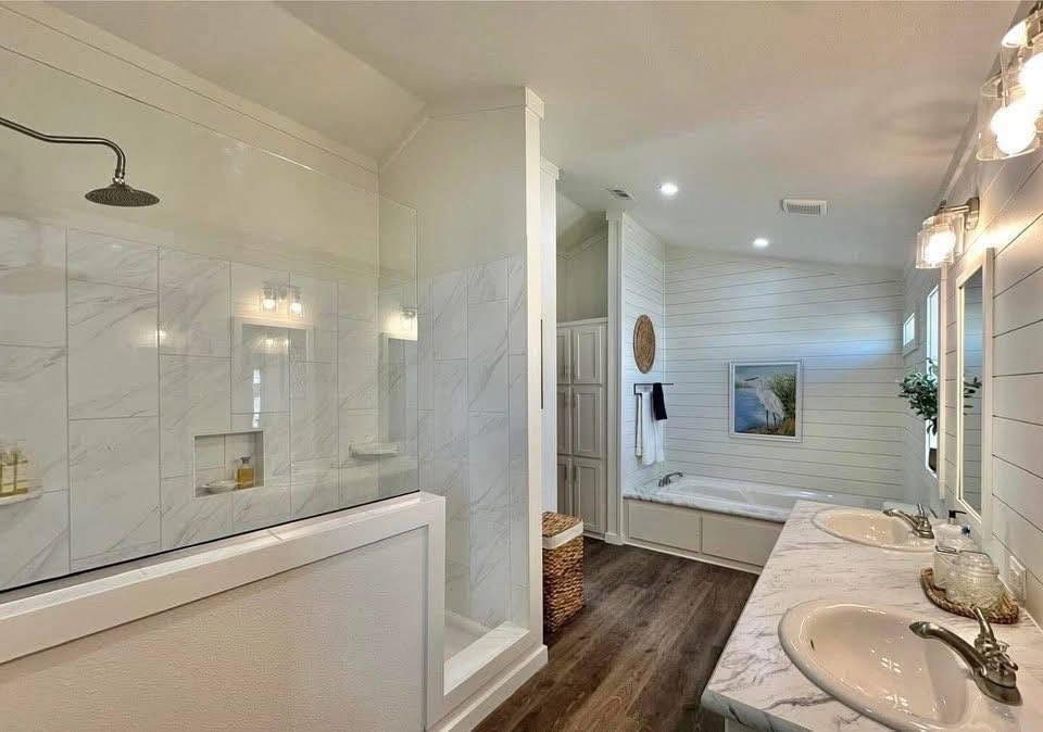 Spacious bathroom with a marble countertop, dual sinks, large shower, and soaking tub. Soft lighting and wood floors create a serene ambiance.