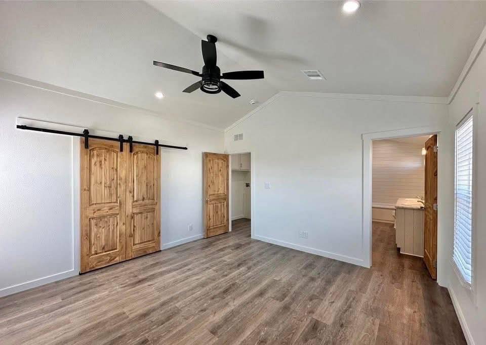 Spacious room with wood floors and white walls. Features sliding wooden barn doors, a ceiling fan, and a doorway leading to another room, creating a warm, inviting atmosphere.