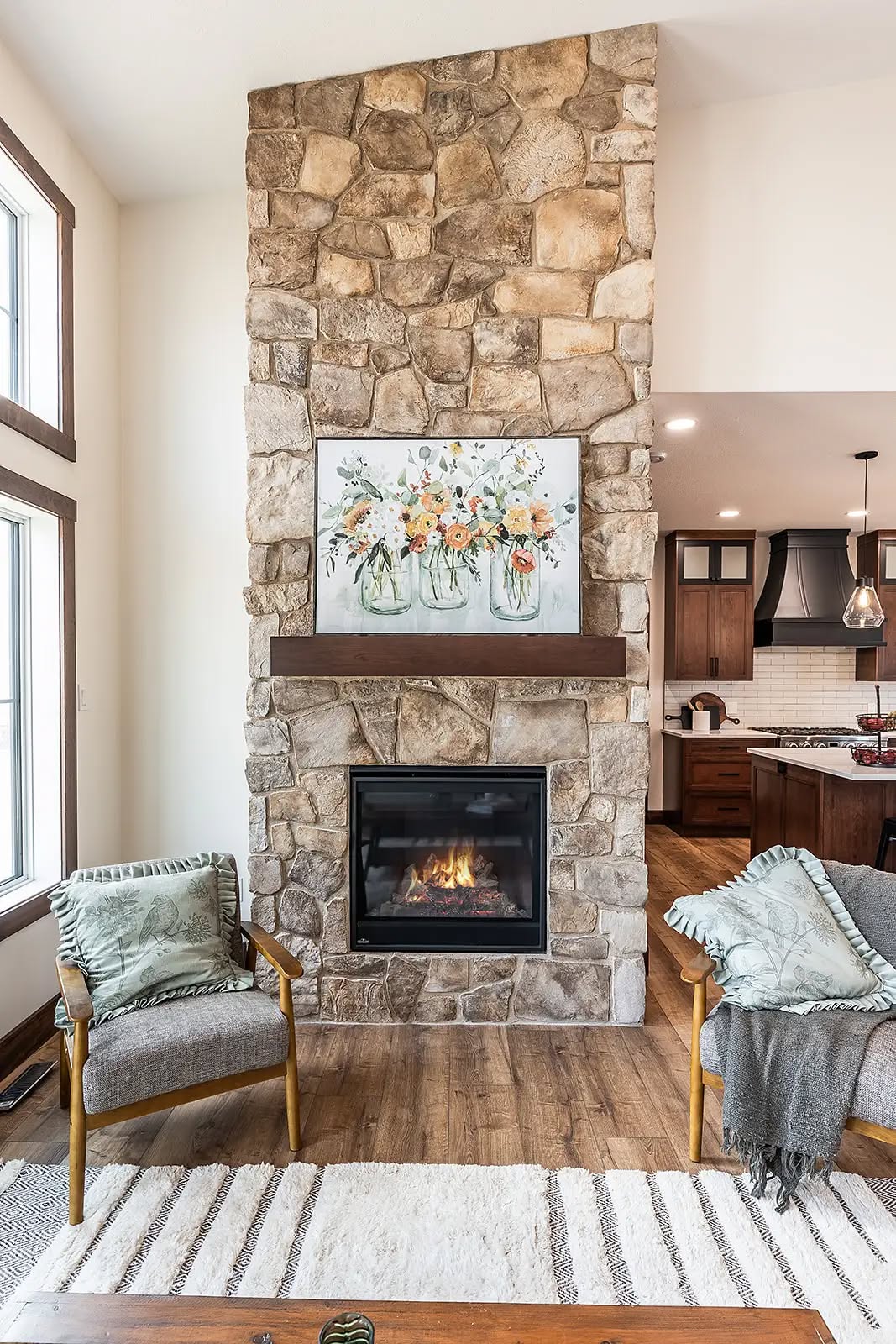 Cozy living room with a stone fireplace featuring a floral painting above it. Two chairs with cushions flank the fireplace, and a kitchen is visible in the background.