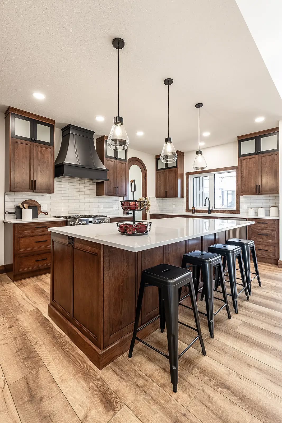 Modern kitchen with wooden cabinets, black range hood, and a large island featuring a white countertop. Hanging lights illuminate, with stools lined up. Cozy and elegant.