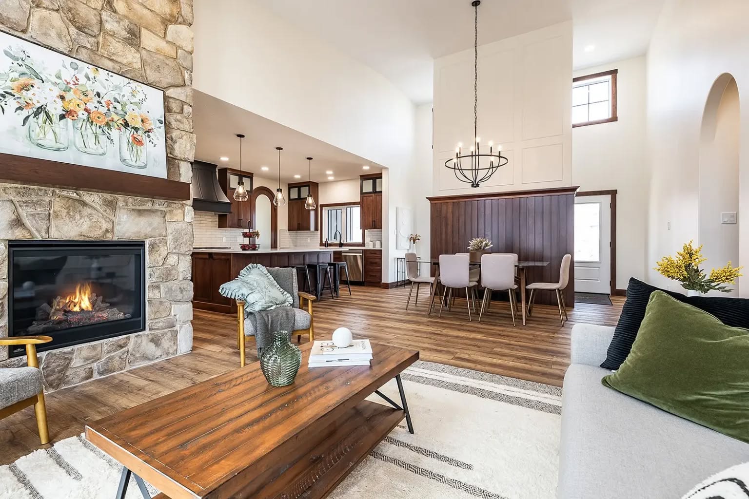 Spacious modern living room with high ceilings, stone fireplace, and cozy furniture. Open dining area with a wooden table and elegant chandelier. Warm, inviting tones.