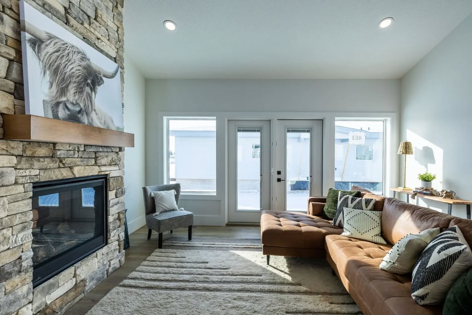 Modern living room with stone fireplace and animal artwork. Brown leather sectional with patterned pillows, large windows, and a cozy chair create a welcoming, bright space.