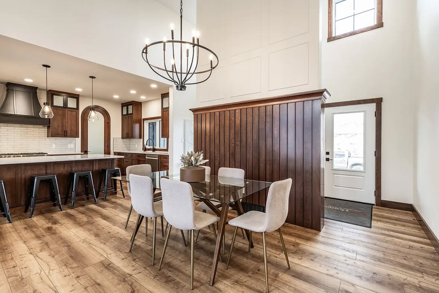Spacious dining area with a glass table, six cream chairs, and a modern chandelier. To the left, a kitchen with wood accents and bar seating. Warm, inviting atmosphere.