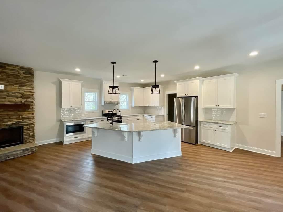 Spacious modern kitchen with white cabinets, marble island, stainless steel appliances, and wood flooring. Pendant lights hang above the island, evoking a bright, inviting atmosphere. A stone fireplace adds warmth.