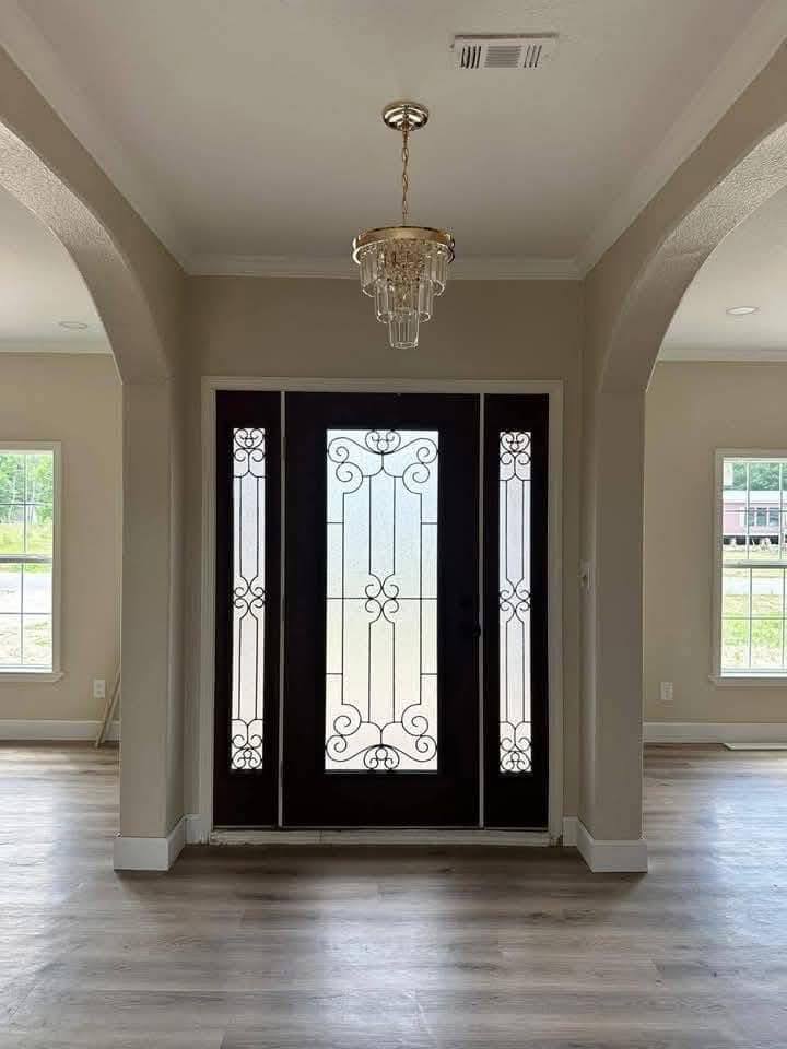 Elegant entryway with a dark wooden front door featuring ornate glass panels. A chandelier hangs overhead, and natural light streams in from side windows.