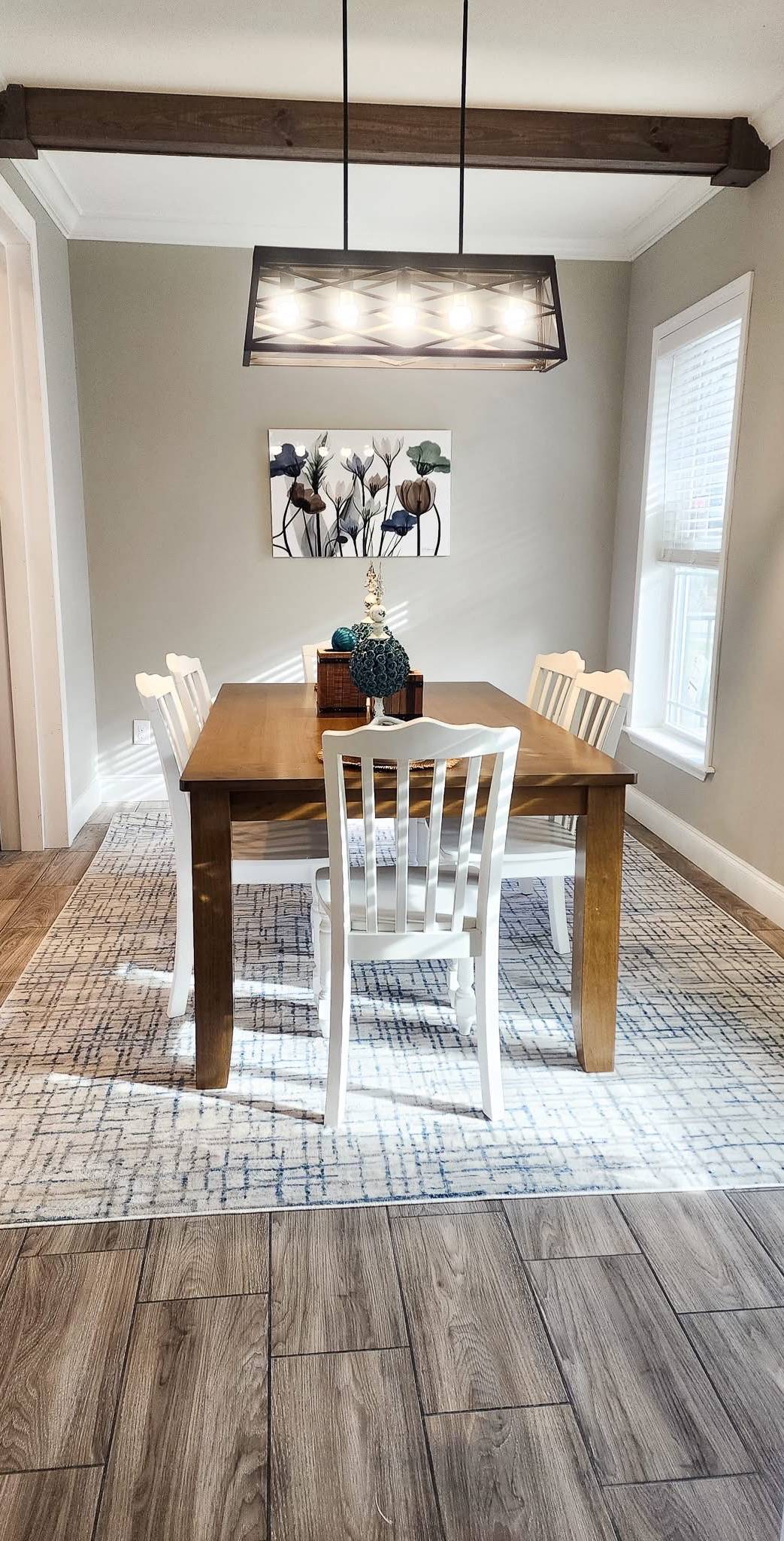 Cozy dining room with a wooden table, four white chairs, and a decorative centerpiece. A floral painting and modern chandelier add elegance.