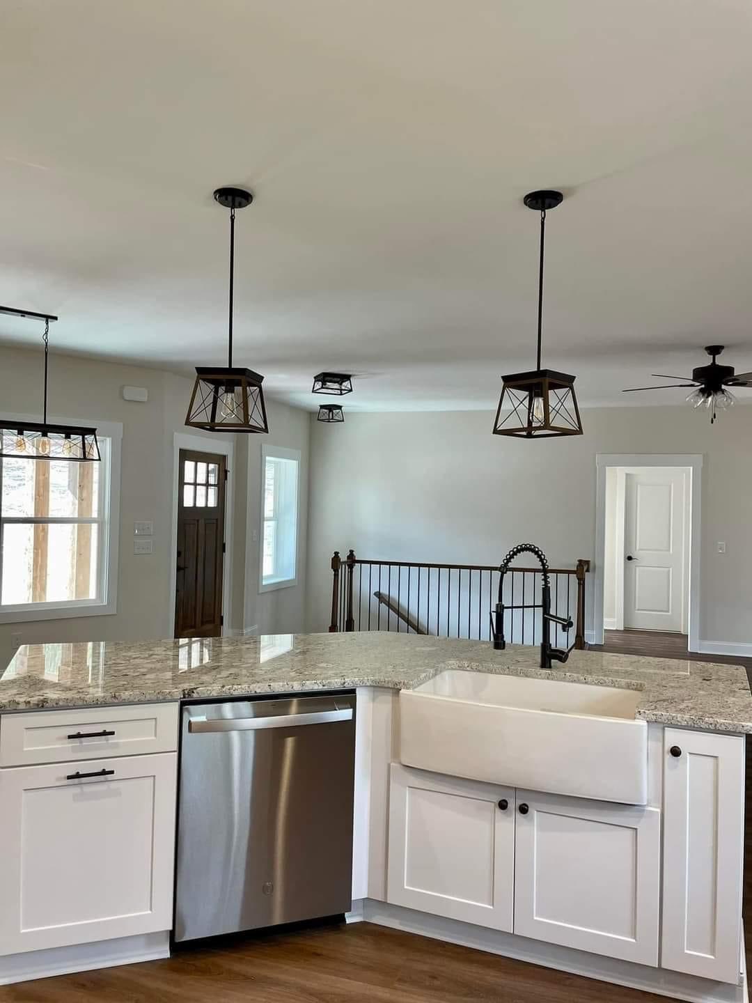 Modern kitchen with white cabinets, granite countertops, and a farmhouse sink. Pendant lights hang overhead. The room has hardwood floors and a visible staircase.