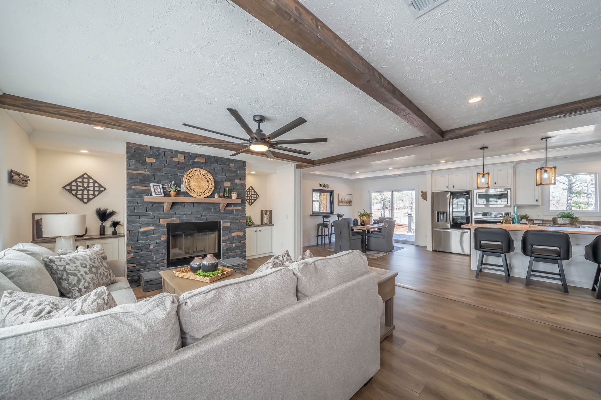 Spacious living room with cozy beige sofas, a modern black stone fireplace, and open kitchen. Wood beams and ceiling fan enhance warmth and style.