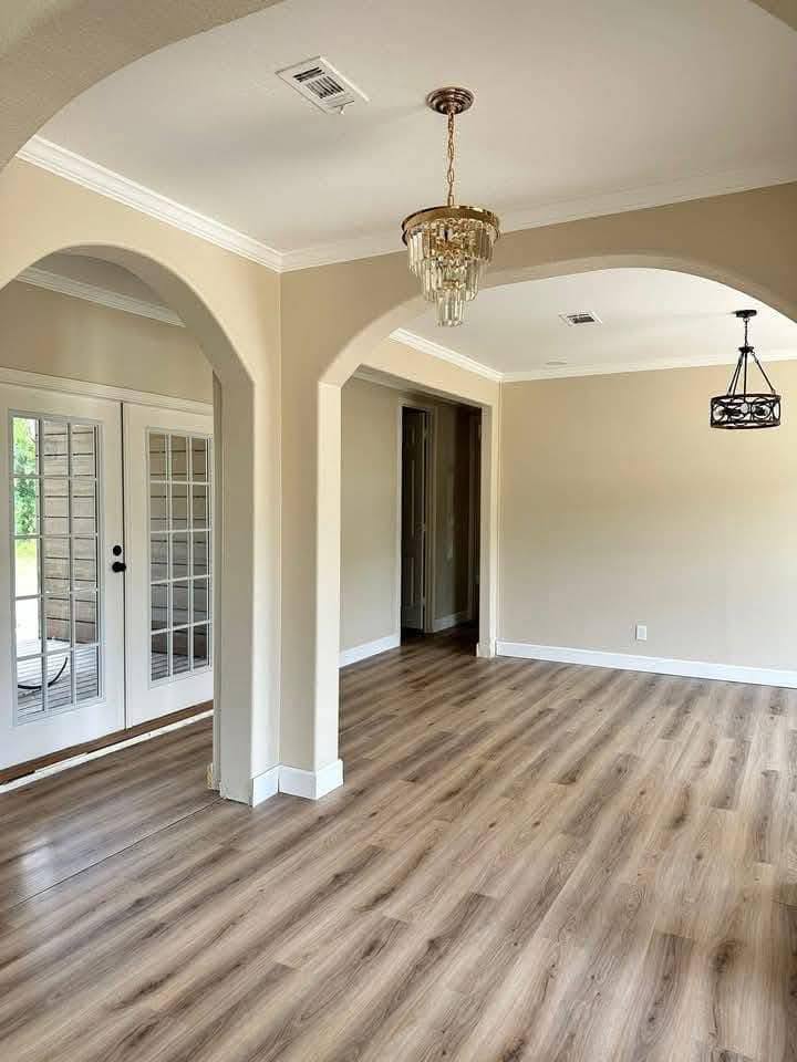 Spacious, empty room with light wood floors, beige walls, and white trim. Arched doorways, glass double doors, and two chandeliers add elegance.