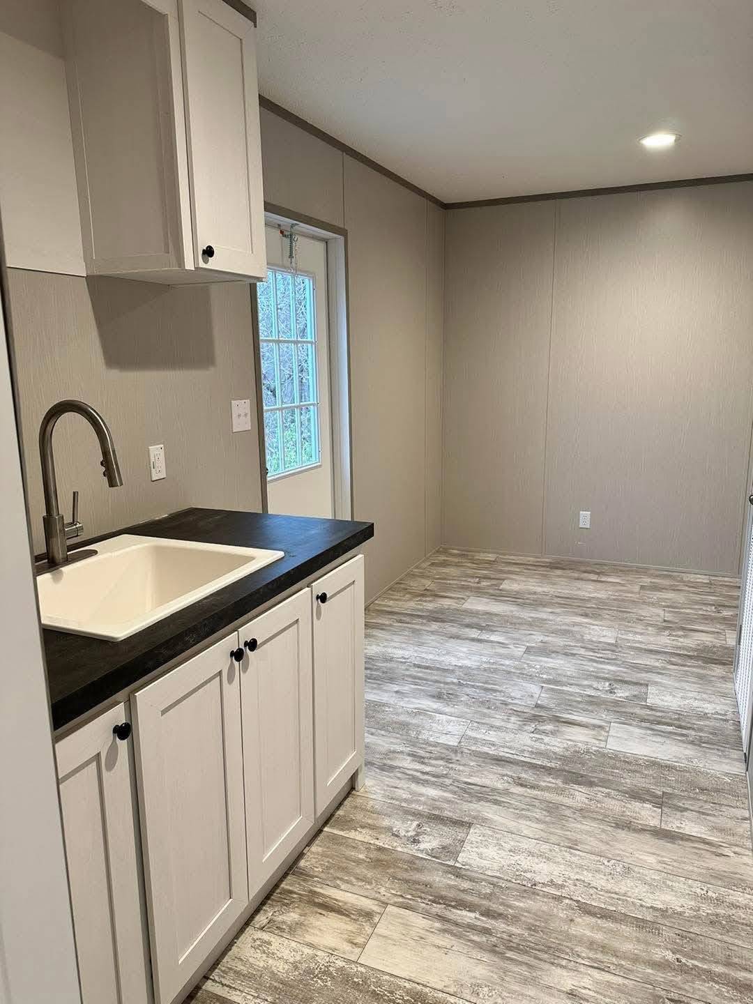 Minimalist kitchen with light gray walls and wood-patterned flooring. Features white cabinets, a black countertop, stainless faucet, and a door with window.
