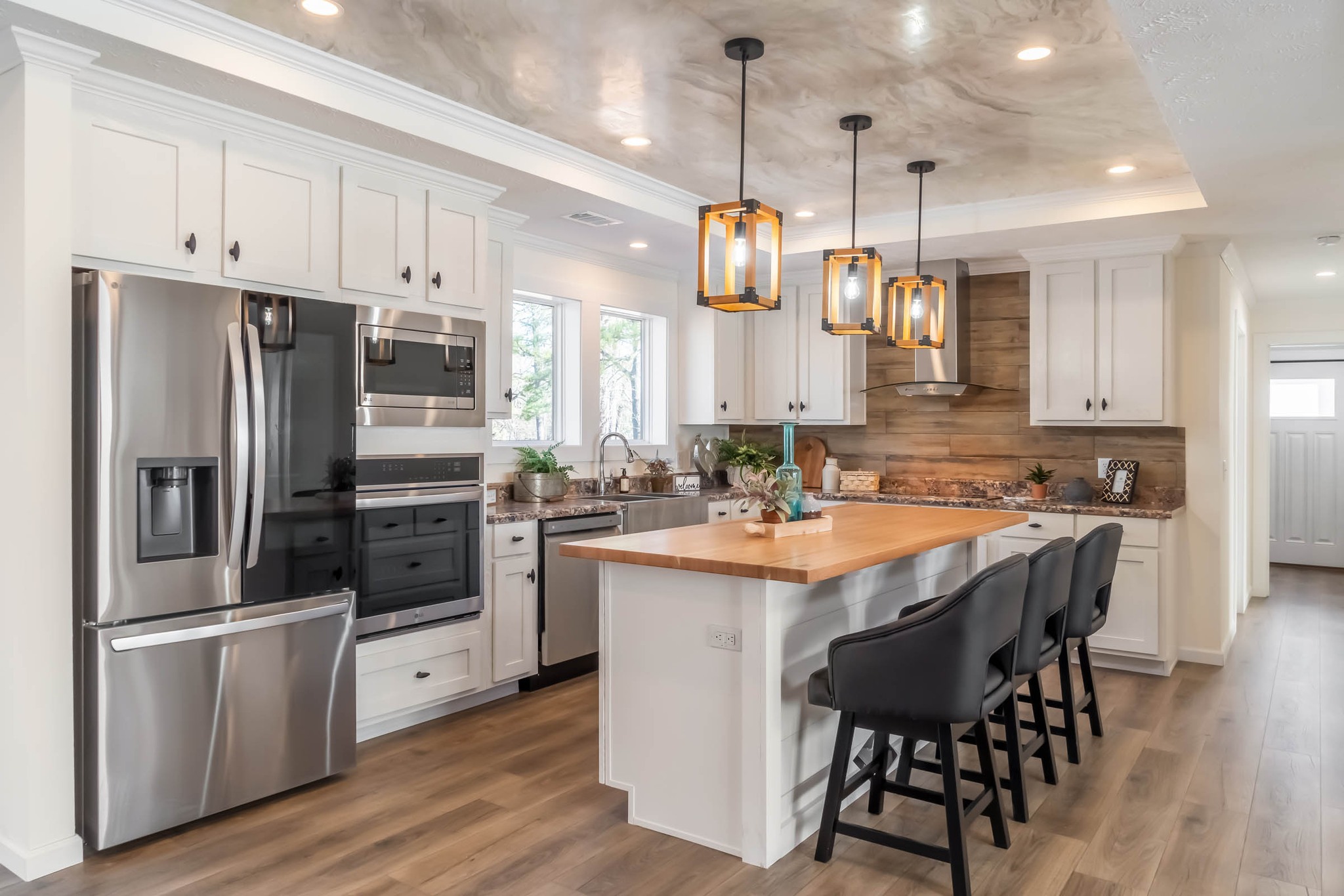 Modern kitchen with sleek stainless steel appliances and white cabinetry. A wooden island with dark chairs is illuminated by stylish pendant lights.