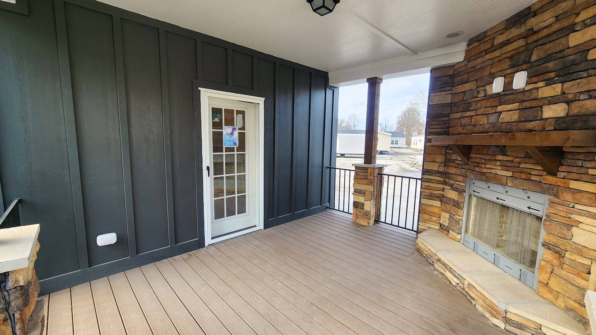 A covered patio features a dark siding wall with a white-framed glass door and a stone fireplace. The space has a cozy and inviting atmosphere.