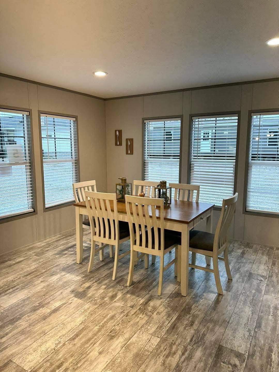 Bright dining room with a wooden table and six chairs on a wooden floor. Large windows with blinds surround the room, creating an airy, inviting space.