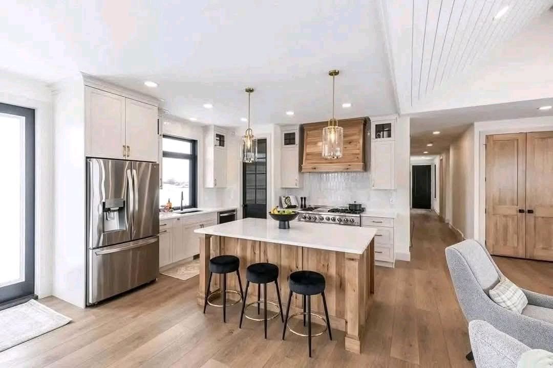 A modern kitchen with light wood floors, white cabinets, and a stainless steel fridge. The island has three black stools and two pendant lights above. Cozy and bright.