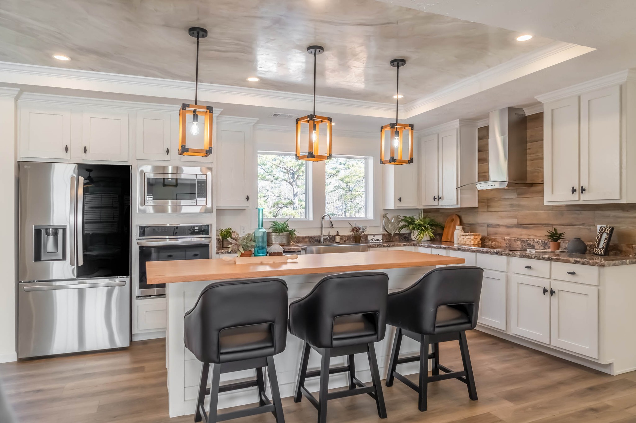 Modern kitchen with white cabinets, stainless steel appliances, and a wooden island with three black chairs. Pendant lights add warmth and elegance.