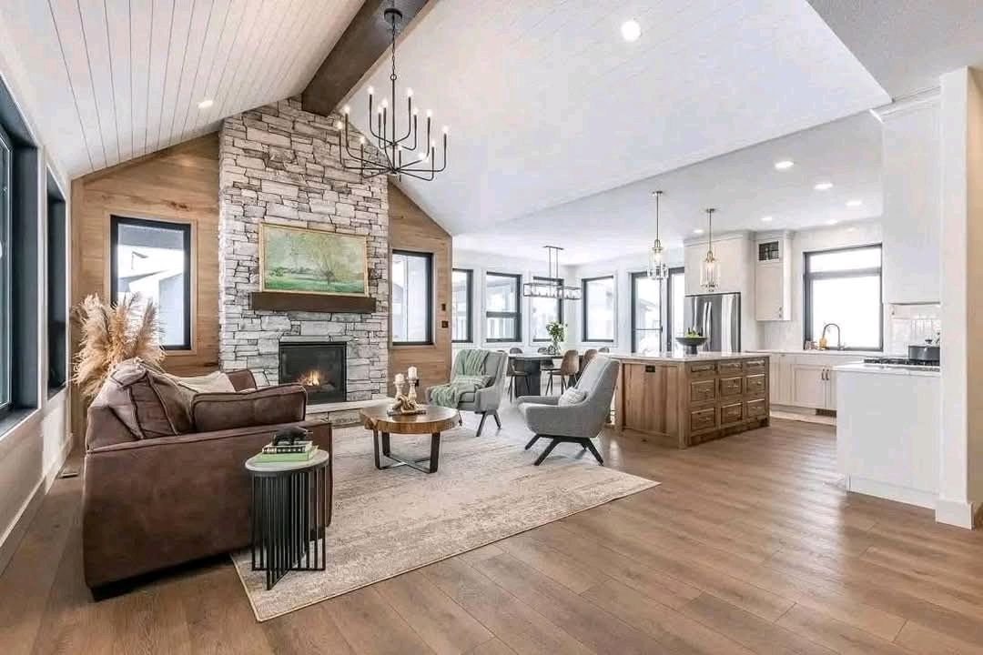 Spacious open-concept living room with high ceilings, a stone fireplace, and modern chandelier. Features a brown sofa, wooden accents, and adjacent kitchen.