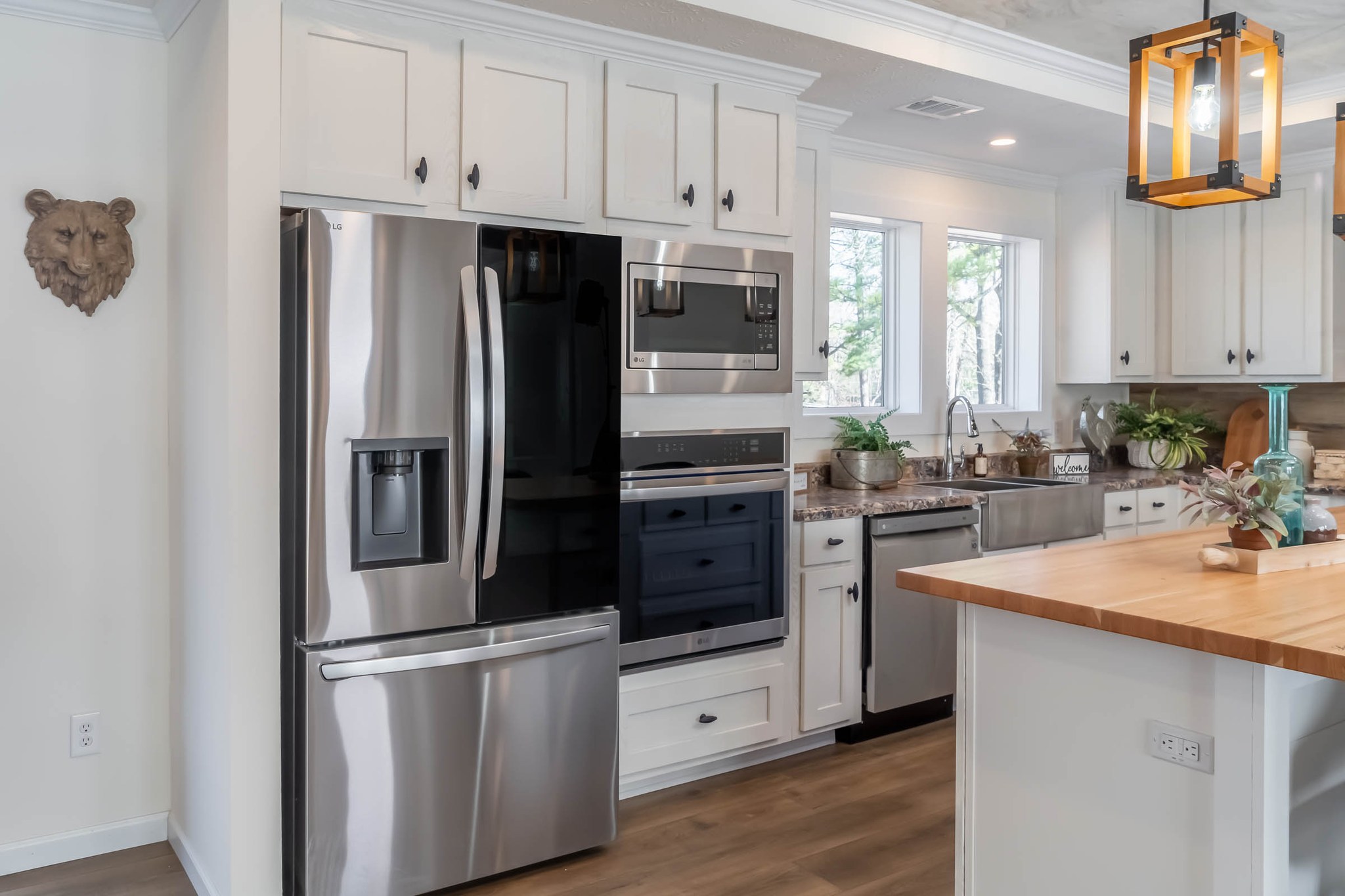 Modern kitchen with stainless steel fridge, oven, and microwave. White cabinets, large window with plants, and a wooden island under a pendant light. Cozy, bright atmosphere.