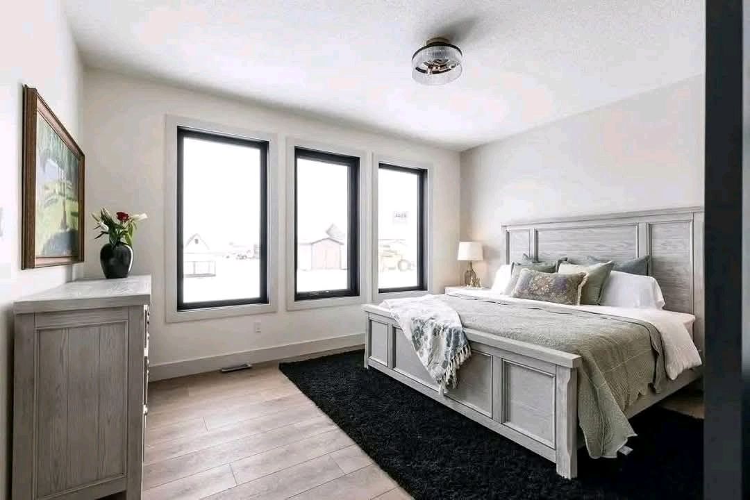 Spacious bedroom with a large bed, light wood furniture, and a black rug. Three tall windows provide natural light. The room feels cozy and serene.