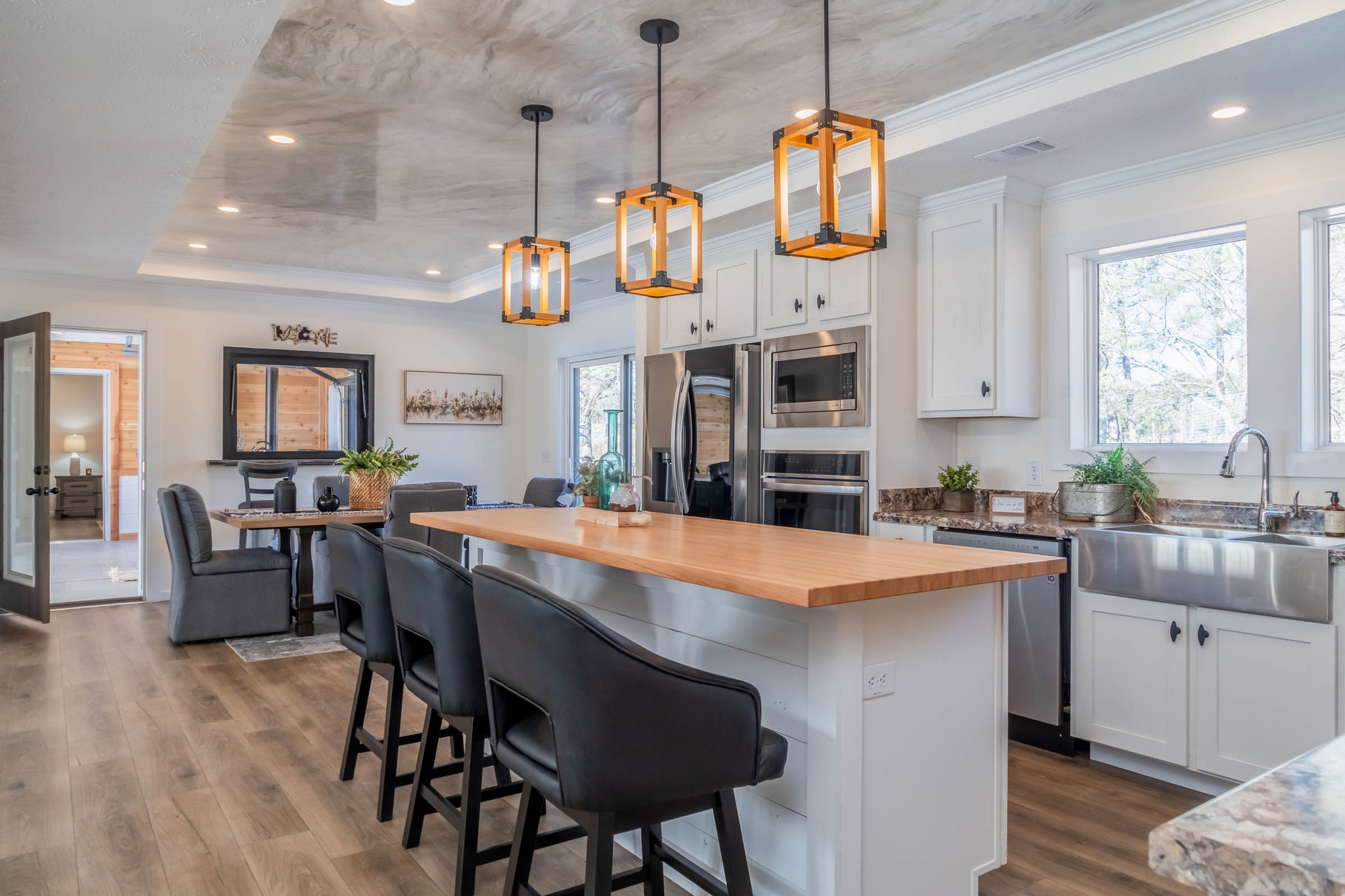 Modern kitchen with a wooden island, three black chairs, pendant lights, and white cabinets. Stainless steel appliances and a farmhouse sink complete the look. Cozy and inviting atmosphere.