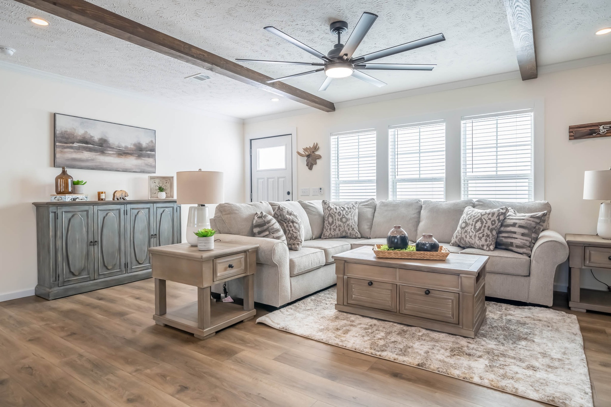 Bright living room with beige sectional sofa, patterned cushions, and a rustic wooden coffee table. Ceiling fan and large windows enhance the airy feel.