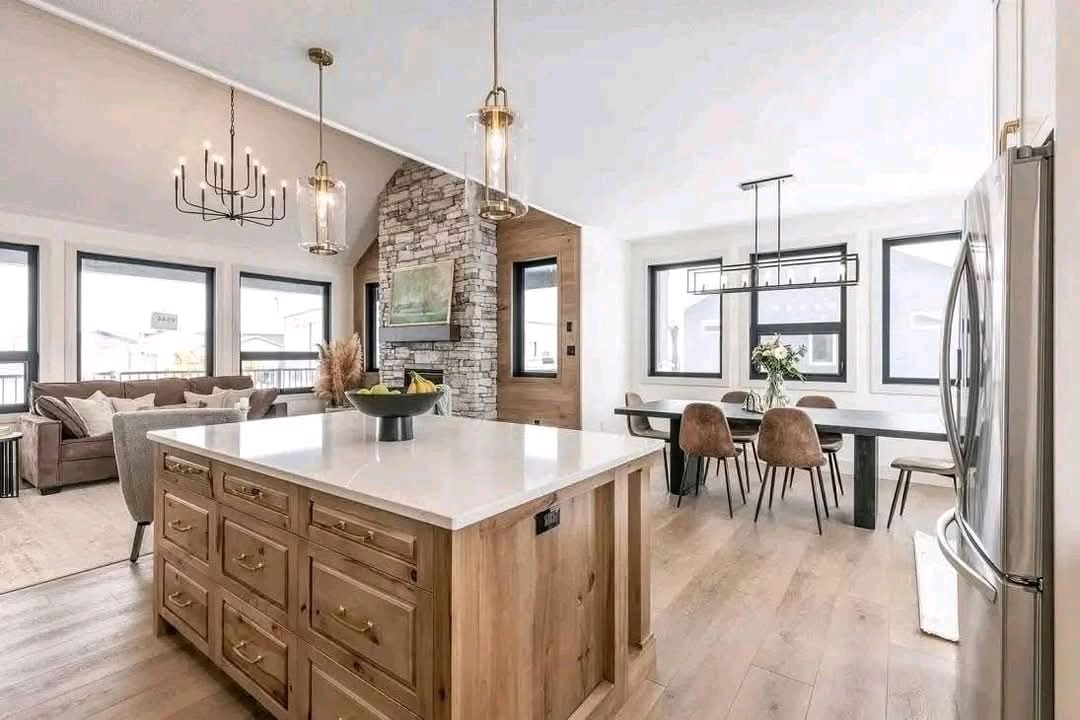 Bright, modern open-plan kitchen and living area with wood accents. Features a central island, stone fireplace, dining area, and cozy seating, exuding warmth.