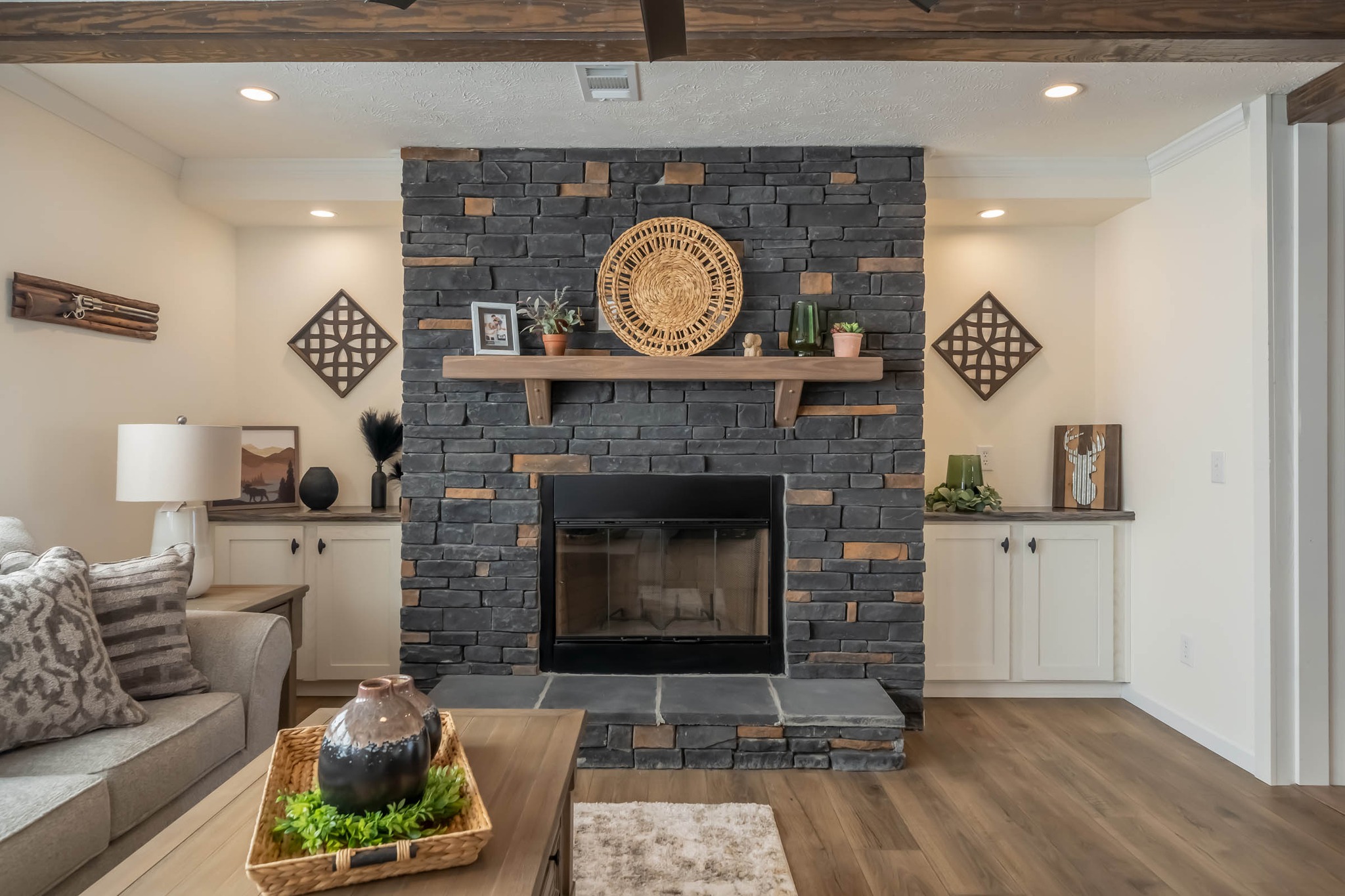 A cozy living room features a dark stone fireplace with a wooden mantel adorned with plants and art. Flanked by white cabinets, it exudes rustic warmth.