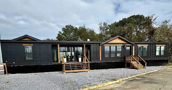 Black modular home with wooden accents, set against a gravel yard and trees in the background. The sky is overcast, creating a calm, rustic feel.