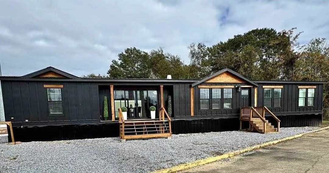 A modern black modular home with wooden accents sits on a gravel lot. There are two wooden staircases leading to glass doors and windows. The sky is overcast.